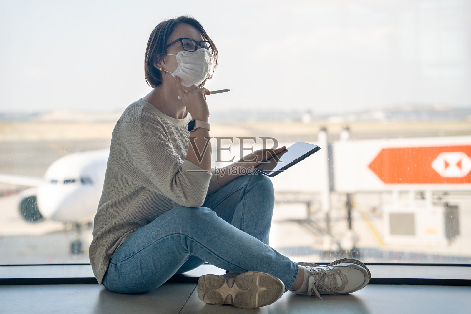 女自由职业者戴着保护面具和眼镜，坐在现代机场靠近窗户的地板上用铅笔在速写本上写东西照片摄影图片
