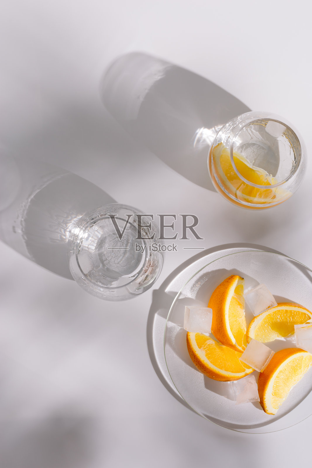 用漂亮的玻璃杯喝纯天然的水和冰，在白色背景的盘子上放一片新鲜的橙子。夏天最小的平坦铺着坚硬的阴影。照片摄影图片
