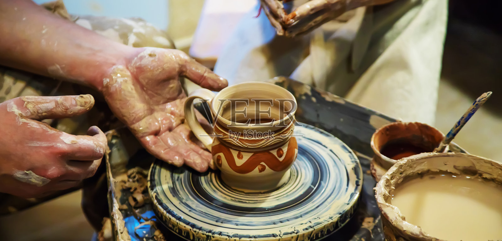 一个师傅和一个学生的手在一个黄泥轮上做一个陶罐。有选择性的重点照片摄影图片