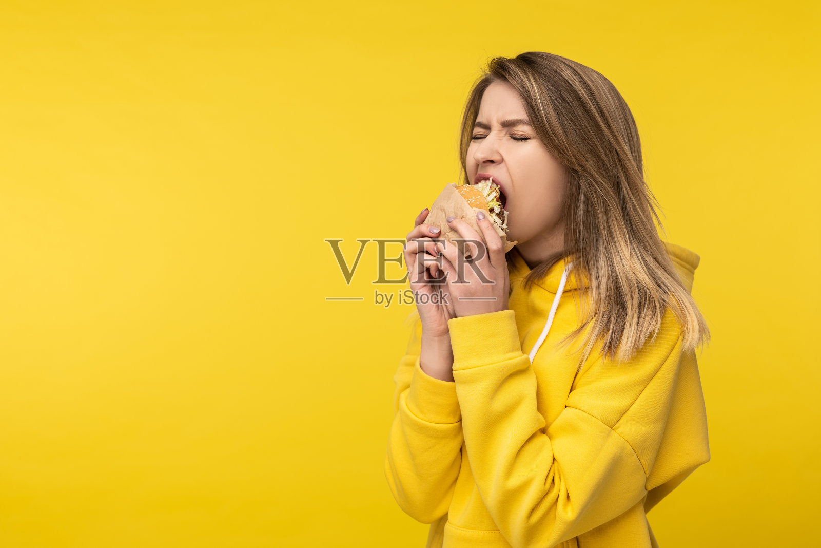 照片上有吸引力的女士在一个强有力的方式咬汉堡。穿着休闲黄色连帽衫，孤立黄色背景照片摄影图片