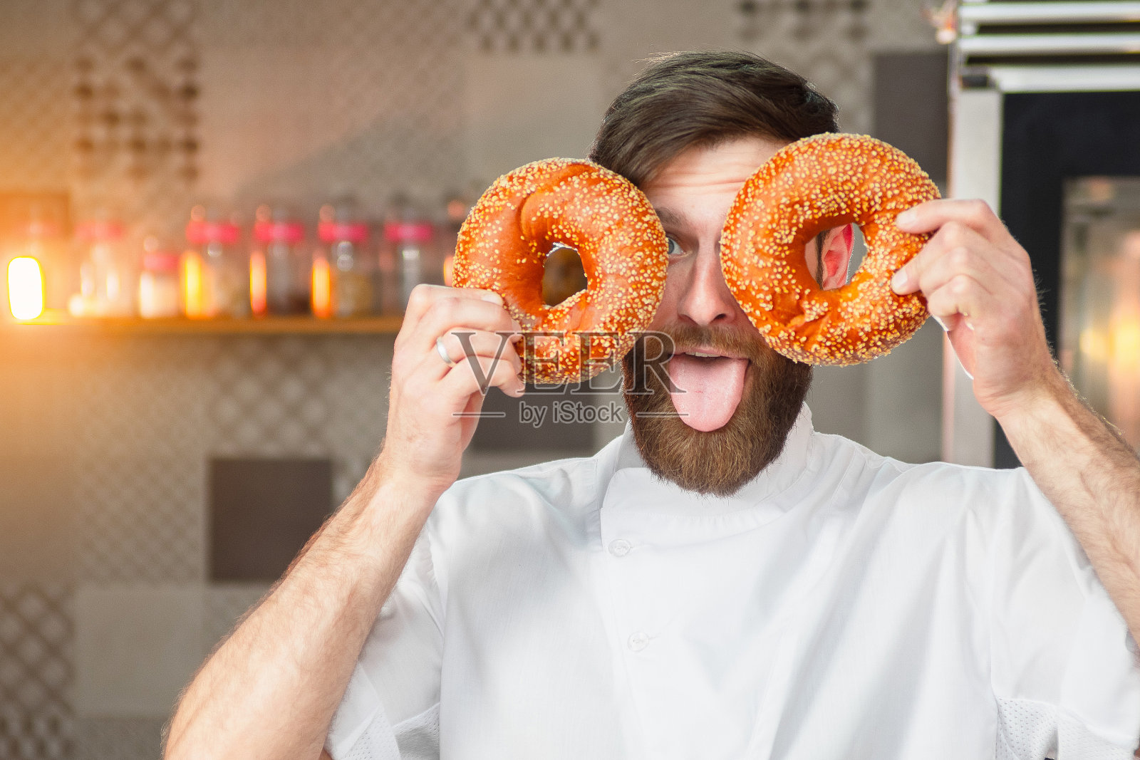 一位年轻英俊的面包师在烤炉的背景上展示了一个笑脸，上面是新鲜的百吉饼，上面有芝麻。照片摄影图片