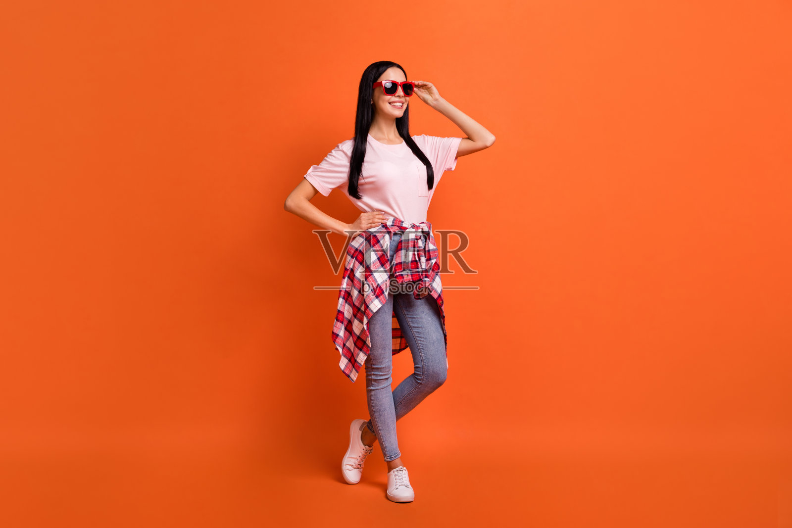全身像的漂亮女孩把手臂放在腰部触摸太阳眼镜微笑孤立的橙色背景照片摄影图片