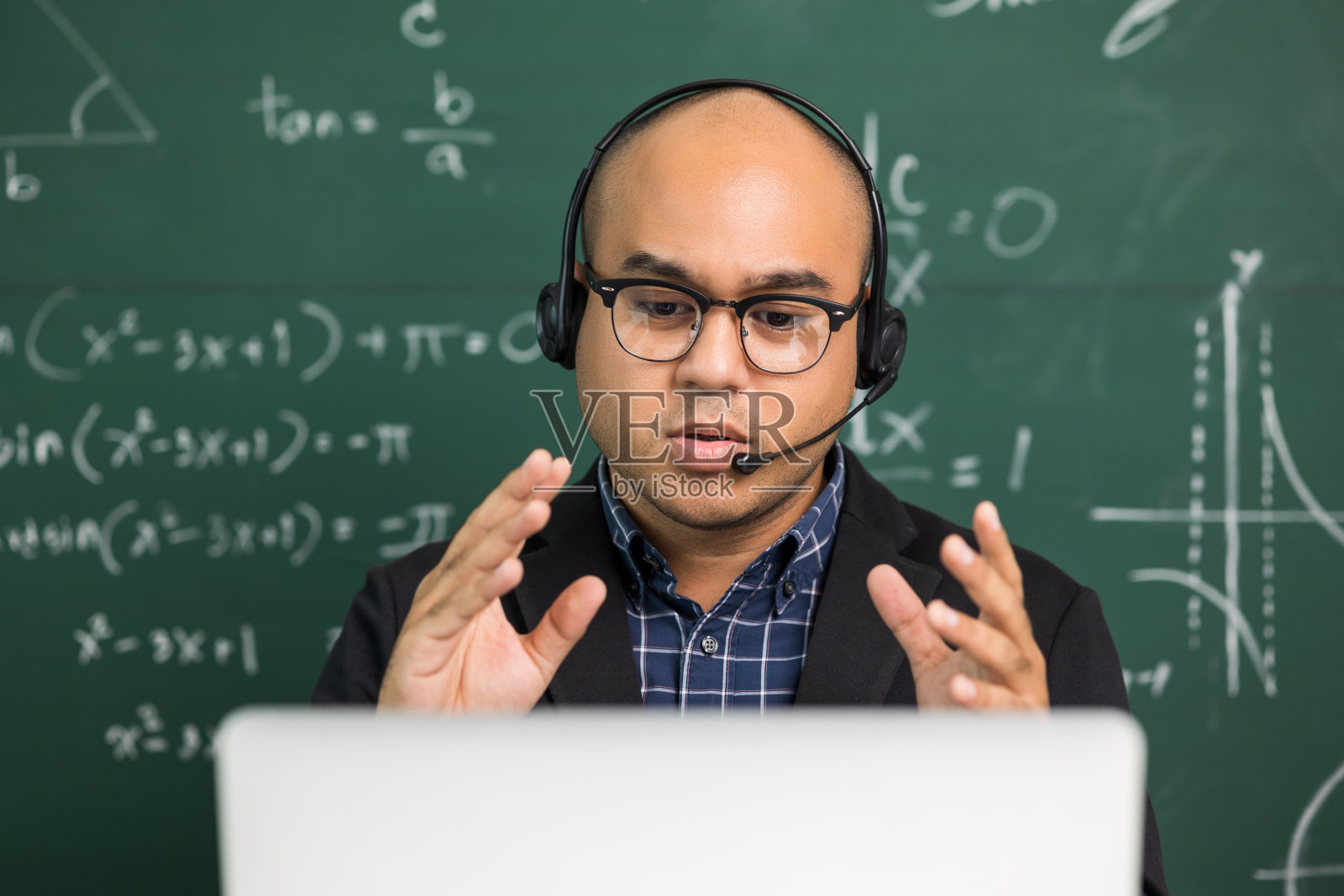 印度年轻教师男子戴着耳机坐着用笔记本电脑进行在线视频会议直播。亚洲数学教师在线授课，供学生在线学习。照片摄影图片