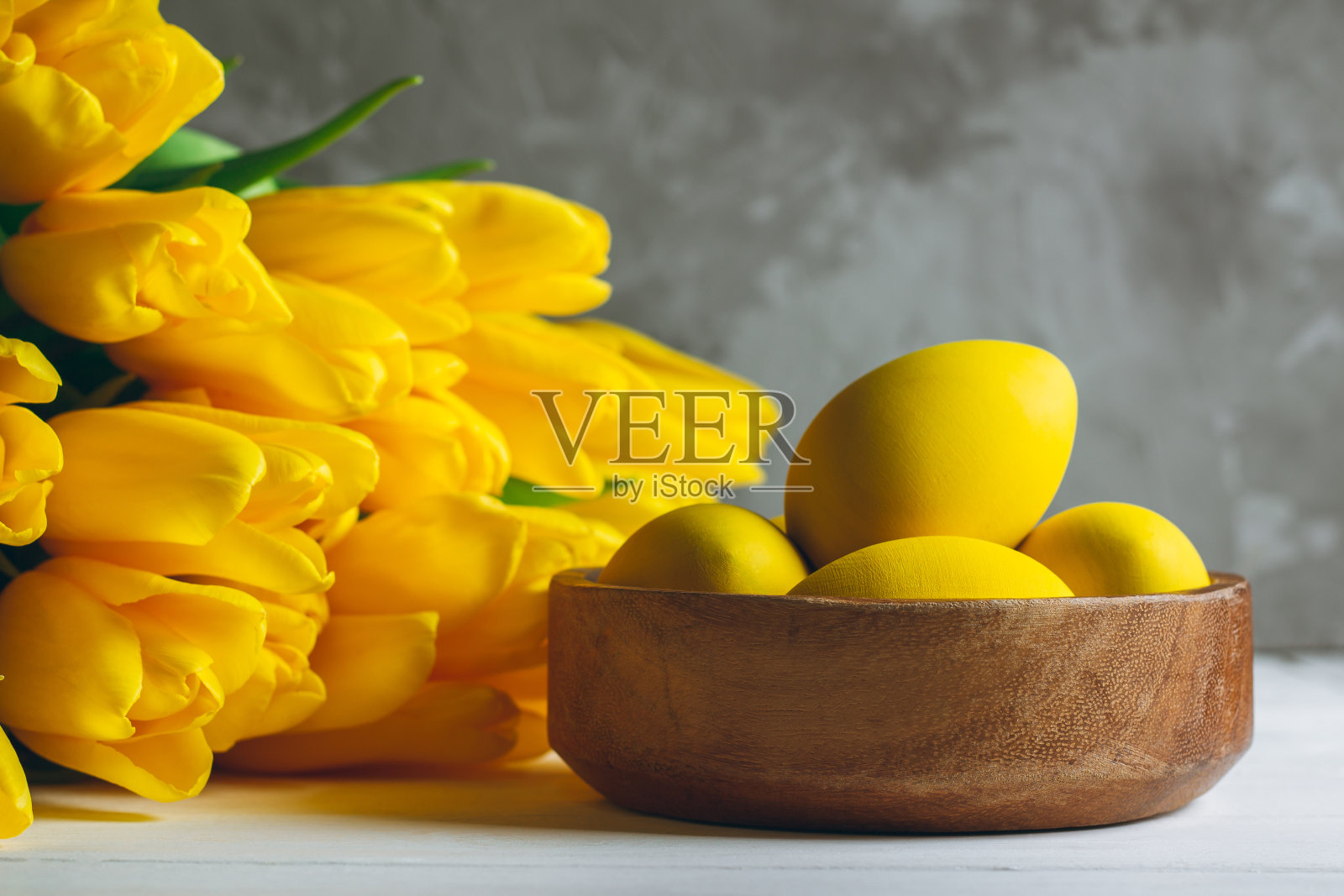 木制的碗里放着复活节彩蛋，白色的木头表面上放着一束亮黄色的郁金香，背景是灰色。本空间照片摄影图片