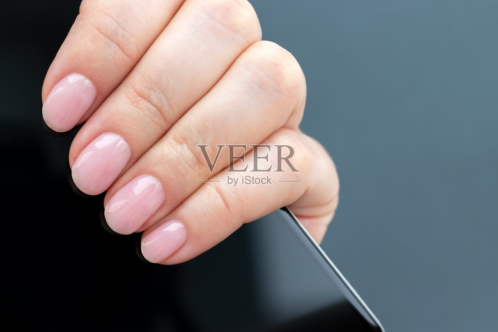 女性手与美丽的美甲-粉色裸指甲与智能手机在灰色背景照片摄影图片