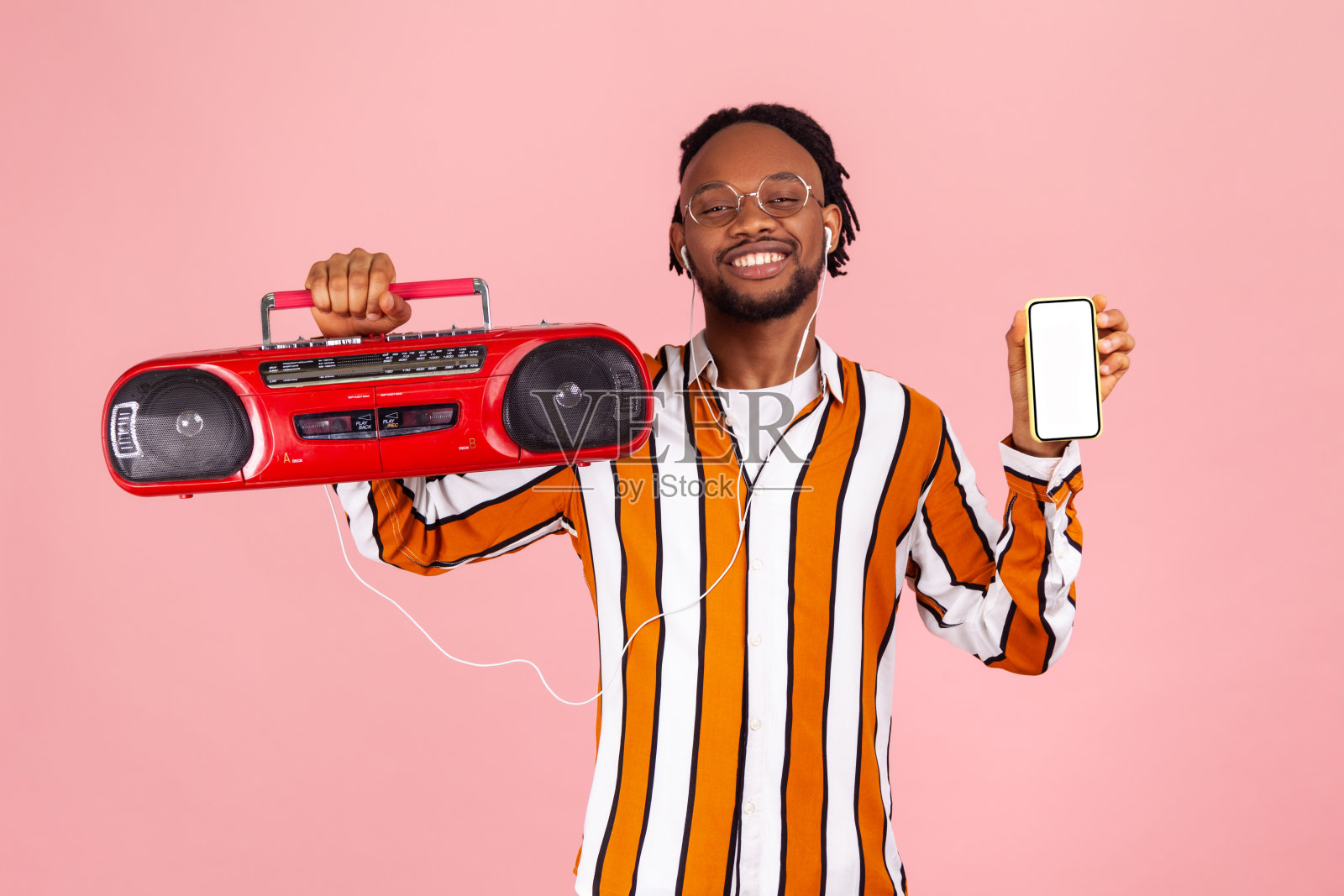 快乐的非裔美国人，梳着雷鬼辫，穿着条纹衬衫，戴着耳机，拿着录音机，用白色屏幕的智能手机寻找音乐应用程序照片摄影图片
