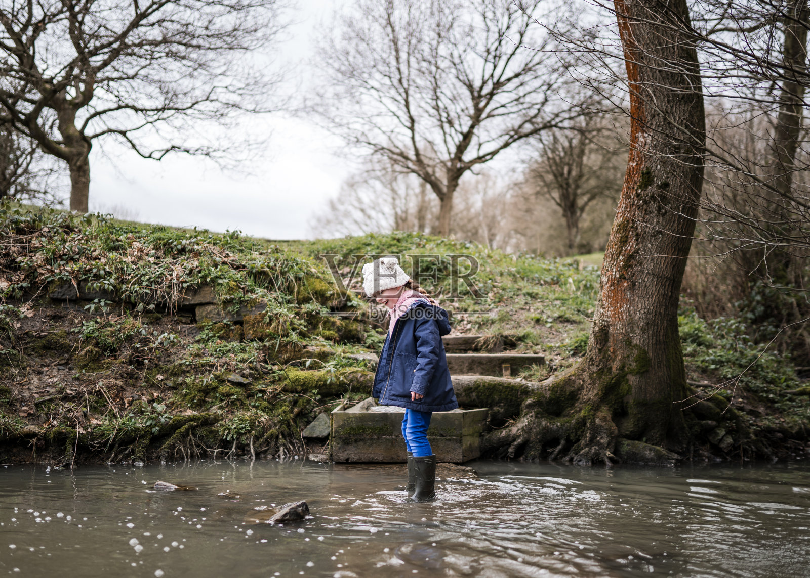 兴奋快乐的年轻女孩穿着惠灵顿靴子走过秋天的乡村，小河溅水，探索伟大的户外自然公园，戴着泡泡帽，穿着蓝色大衣在冬天照片摄影图片