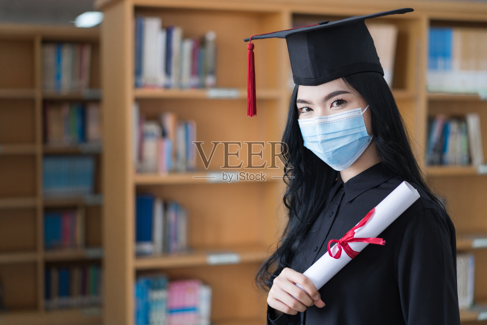 一名年轻快乐的亚洲女子大学毕业生在毕业典礼上穿着毕业礼服和帽子戴着口罩手持和显示学位证书来庆祝她的教育成就照片摄影图片