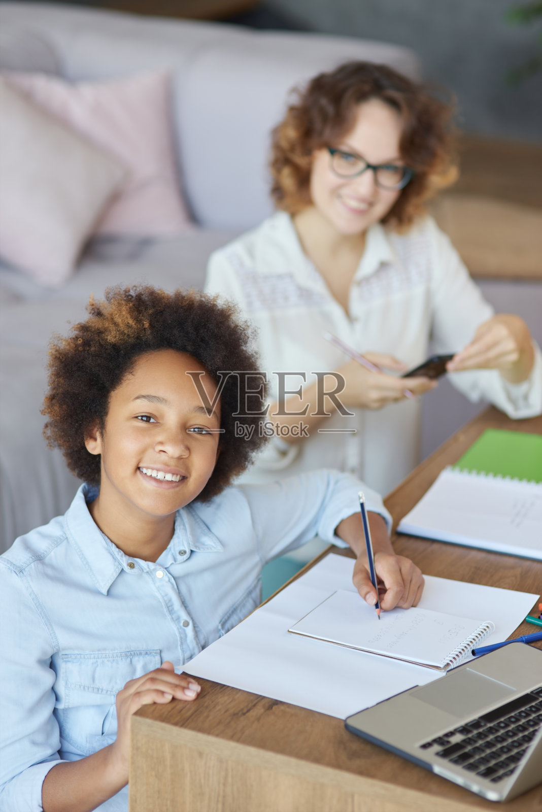 散发出幸福。快乐的混合种族少女在镜头微笑，而有一个女老师上课，坐在家里的桌子照片摄影图片