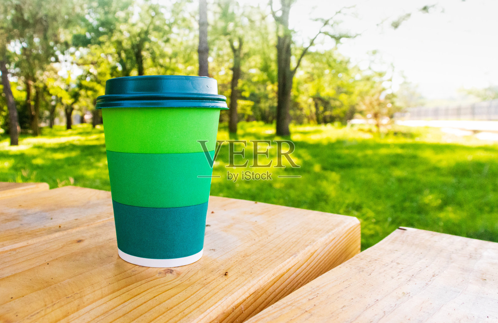 在阳光明媚的天气里，一个带有绿色条纹的纸杯和咖啡架放在木凳上，背景是模糊的美丽绿色。照片摄影图片
