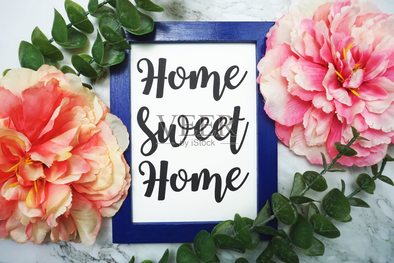 蓝色的画框上写着“甜蜜的家”，绿色的叶子和粉红色的花朵平躺在大理石的背景上照片摄影图片