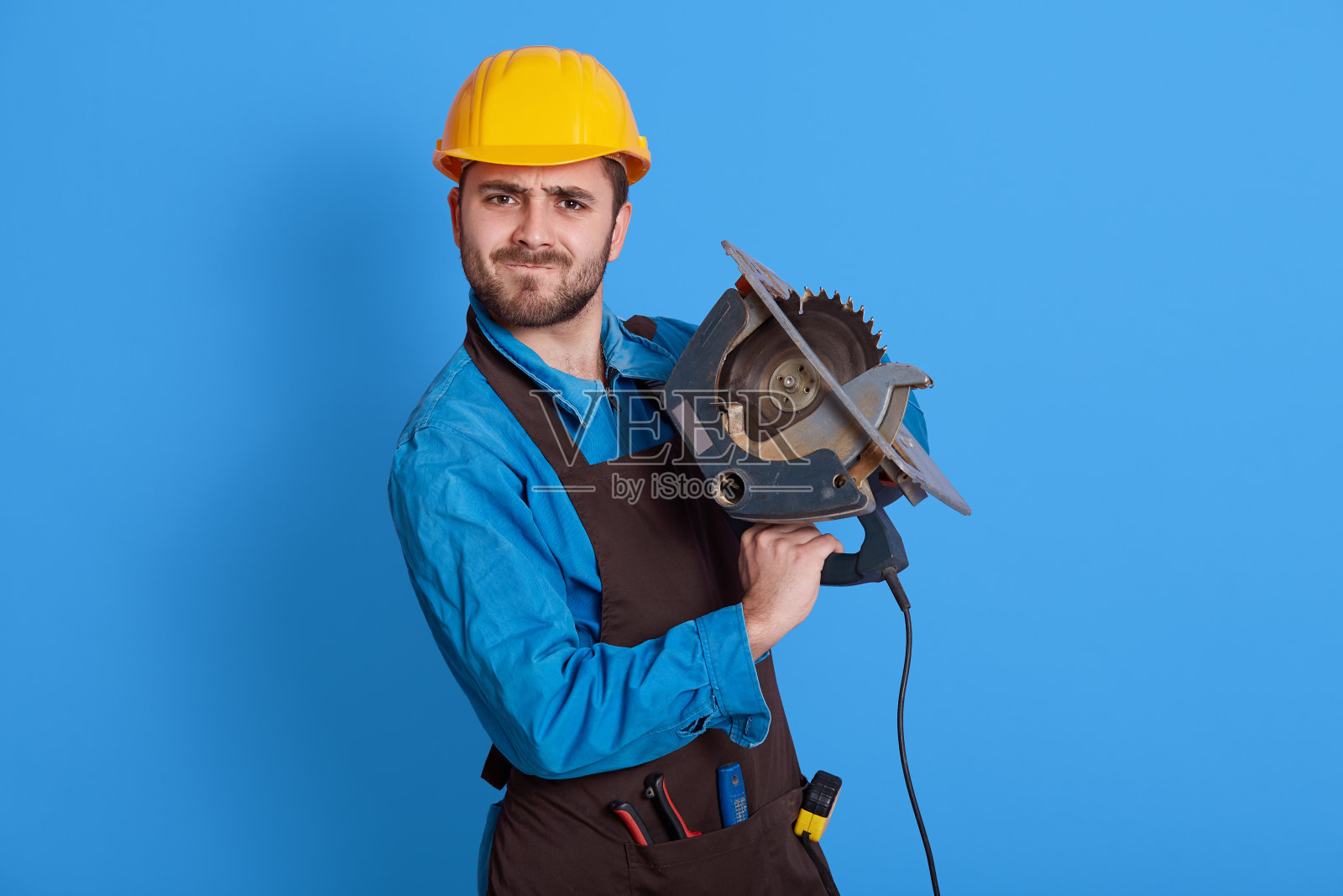 强壮的工人手里拿着圆锯直视着镜头，眉头紧锁，戴着防护头盔和眉毛围裙，在蓝色背景下摆出孤立的姿势。照片摄影图片