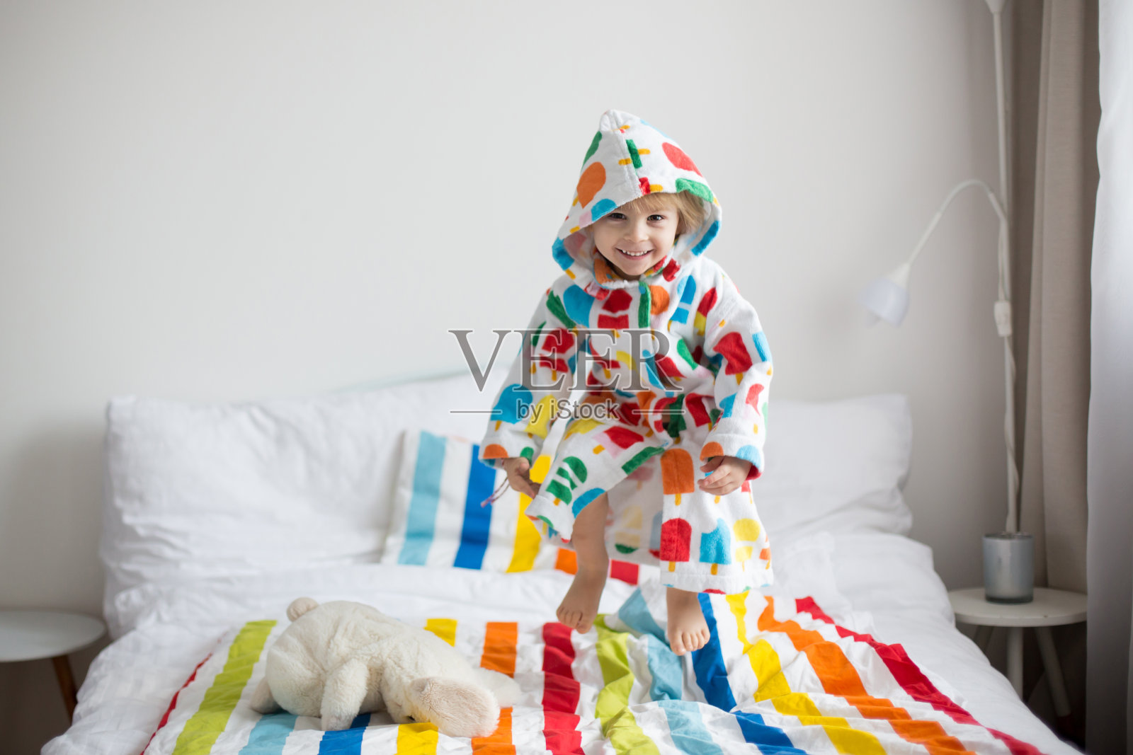 快乐的蹒跚学步的孩子，金发男孩穿着五颜六色的浴袍，在床上跳照片摄影图片