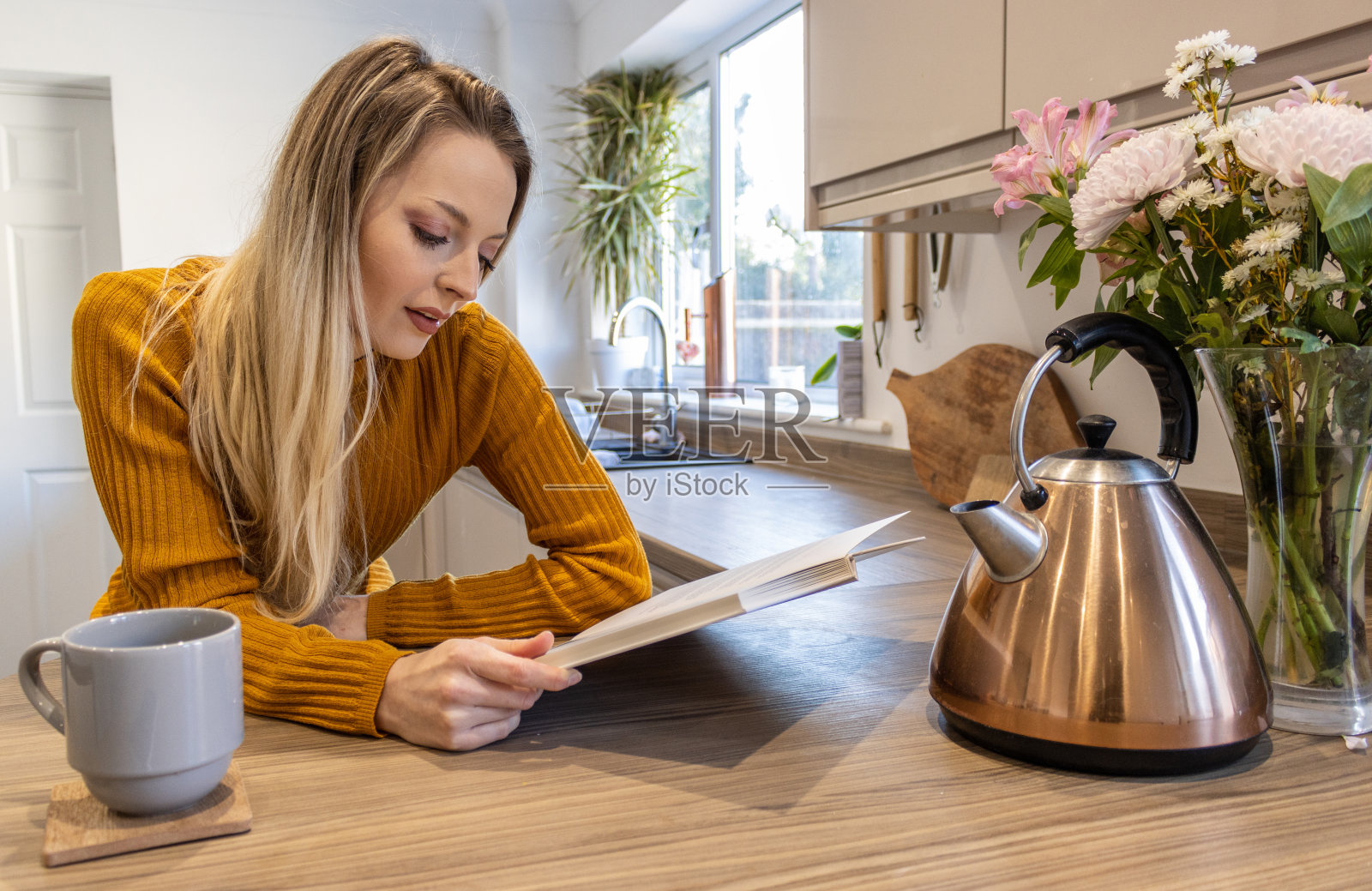 一个金发迷人的年轻学生正斜靠在厨房的柜台上，端着一杯热茶，一边欣赏着一边读着一本书照片摄影图片
