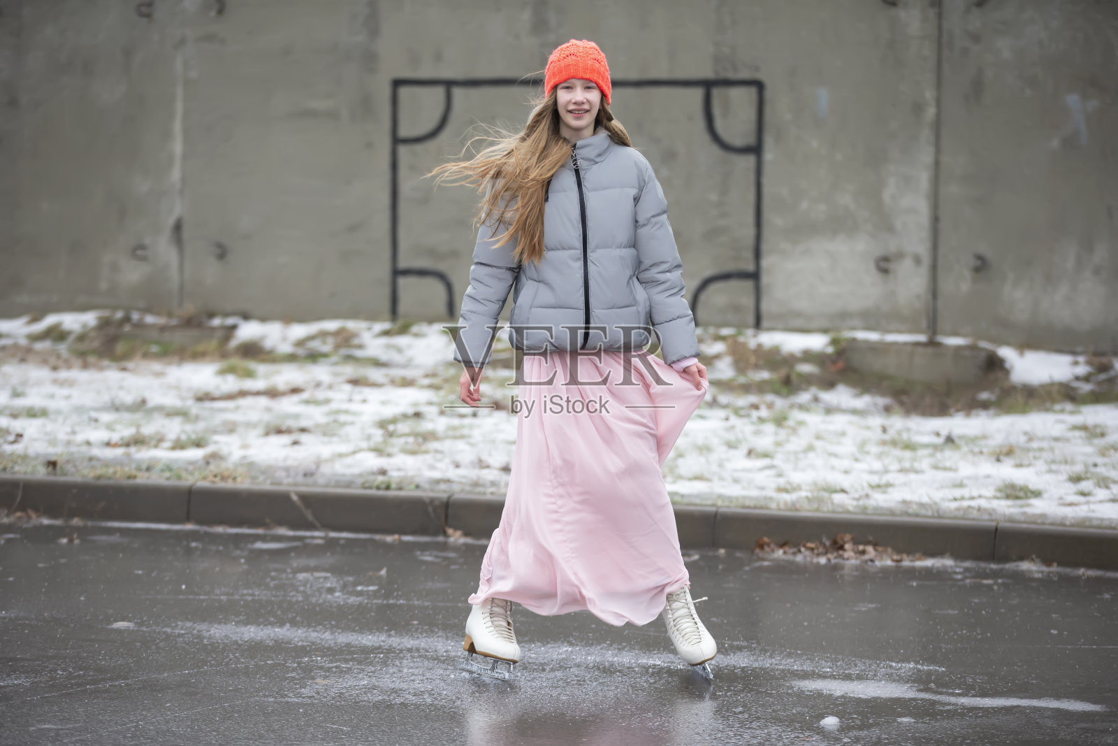 在水泥墙的映衬下，身穿粉色长裙的少女正在冰上滑冰。照片摄影图片