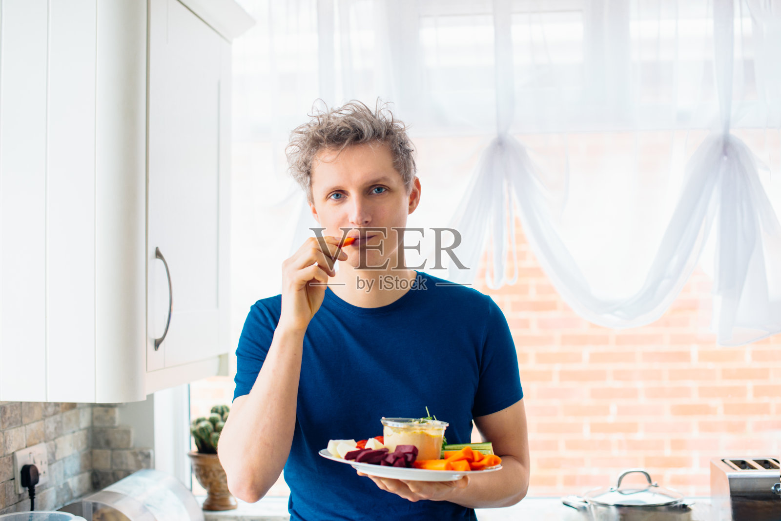 年轻人在厨房里吃着蘸着鹰嘴豆泥的胡萝卜棒。鹰嘴豆泥配生蔬菜放在盘子里。健康食品的午餐。素食和纯素饮食。软选择聚焦。副本的空间。照片摄影图片
