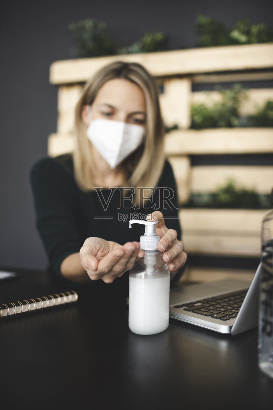 漂亮的，年轻的女商人坐在一个可持续的，生态的办公室戴着一个保护口罩和消毒她的手”n照片摄影图片
