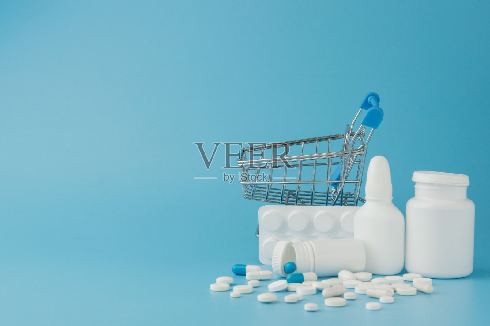 蓝色背景上散落着各种药片、药品、喷雾、瓶子、温度计、注射器和空的购物车。药房购物的概念照片摄影图片