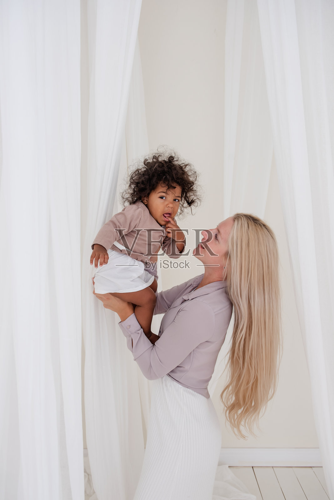 年轻的金发白种人母亲抱着非洲裔美国卷发女儿。在飘动的薄纱的白色剪裁中，女人拥抱女孩亲吻。母亲的爱。领养，混血家庭照片摄影图片