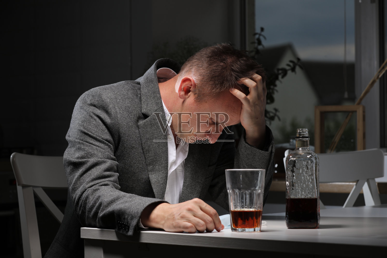 男人下班后在厨房喝酒。患有强烈头痛或偏头痛的年轻人坐在厨房里，喝着一杯威士忌。酒精依赖照片摄影图片