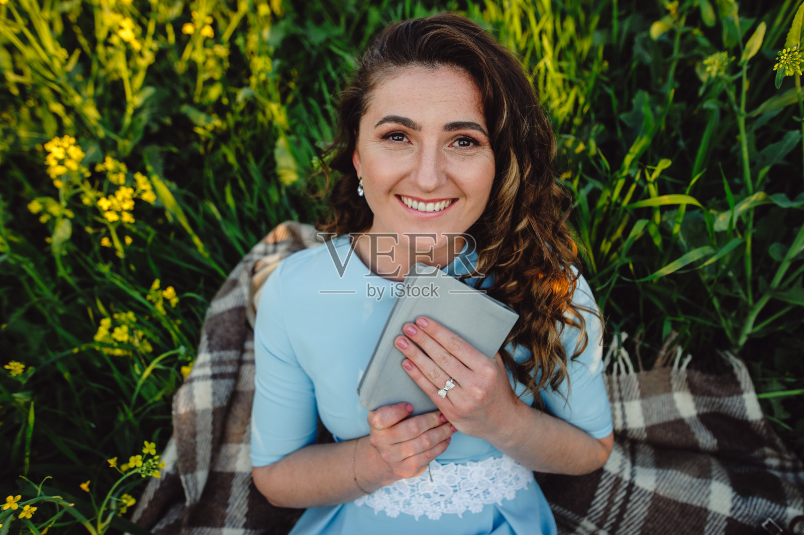 一个穿着蓝色连衣裙的美丽女孩坐在黄花中间的格子布上，手里拿着一本书。一幅以野花为背景的女人肖像。照片摄影图片