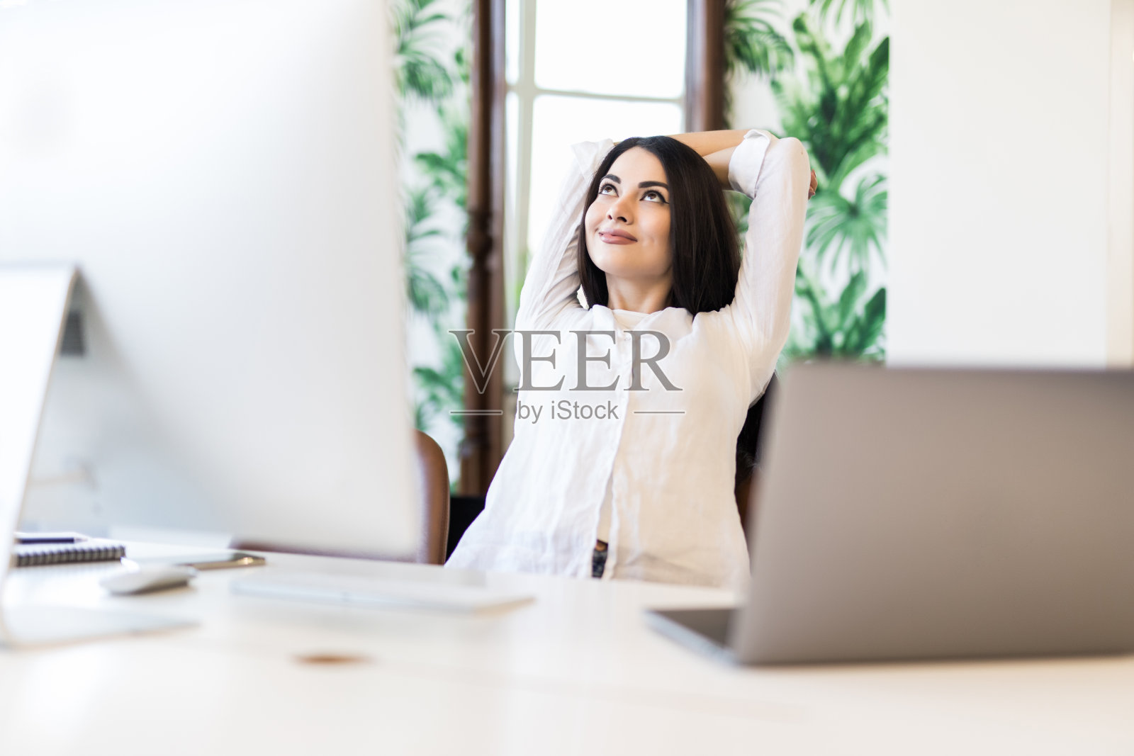 一个快乐的漂亮女人双手放在脑后坐在桌前。女商人闭着眼睛想象着幸福的未来。放松的女孩休息在办公椅。商业成功理念带来的极大愉悦照片摄影图片