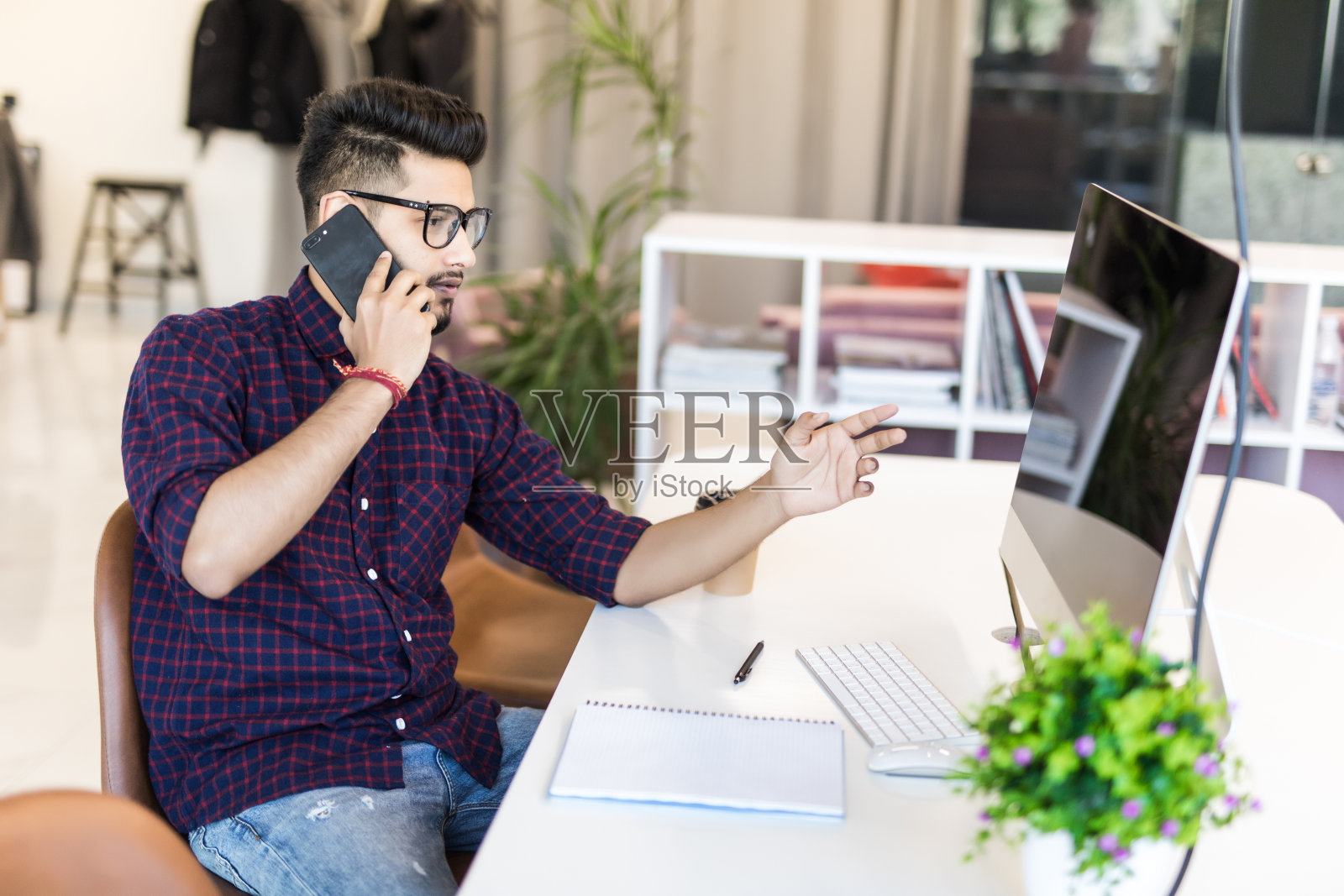 自信的年轻印度男子穿着智能休闲装，拿着智能手机，坐在办公室的工作场所看外面。照片摄影图片