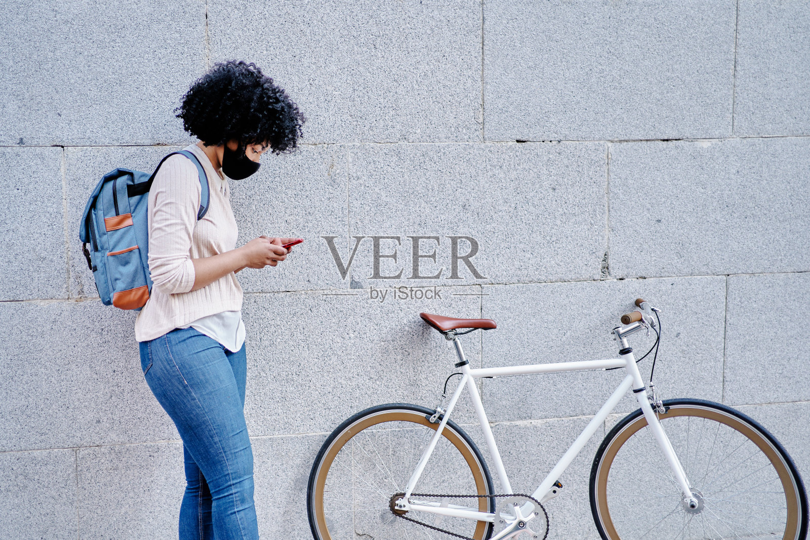 一个手里拿着智能手机的非裔美国女性。戴着防护面具的女人。在城市里骑自行车。照片摄影图片