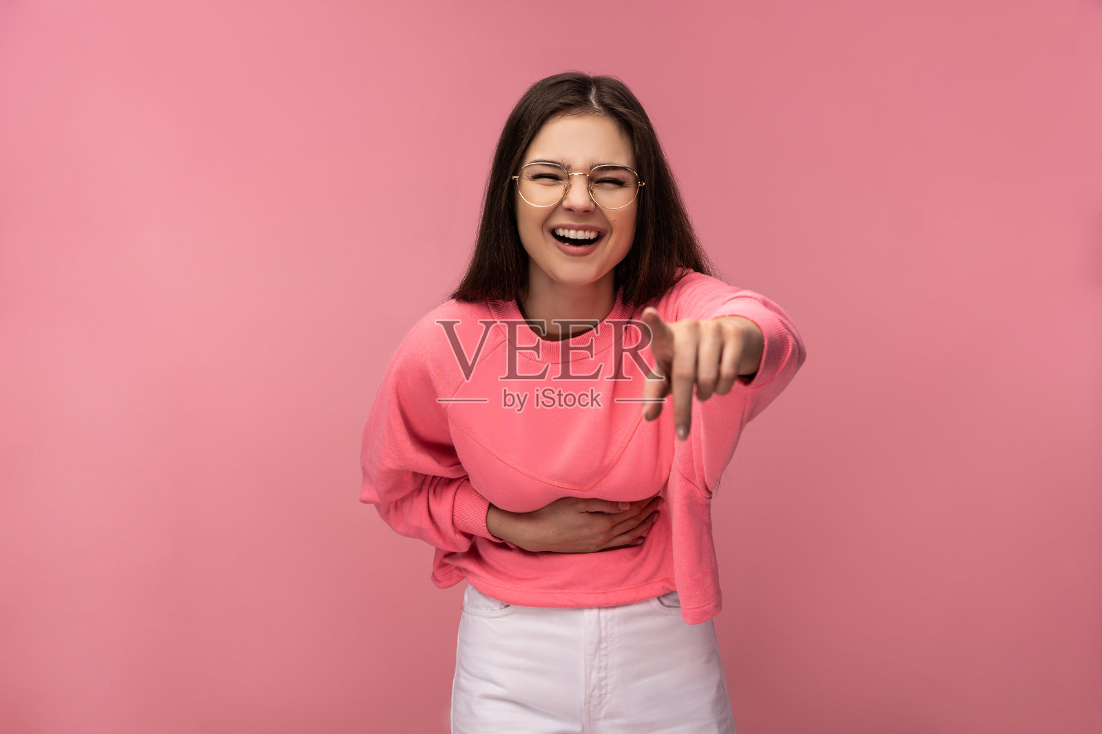 照片中迷人的年轻女子在眼镜指着镜头笑，穿着休闲粉色t恤白色裤子孤立的粉色背景照片摄影图片