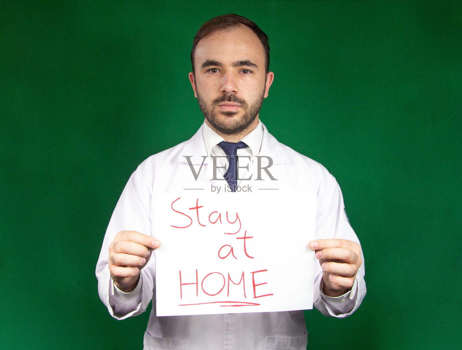 年轻的白种医生，面目全非，举着"待在家里"的牌子。2019冠状病毒病大流行尚未结束，我们必须照顾好自己。照片摄影图片