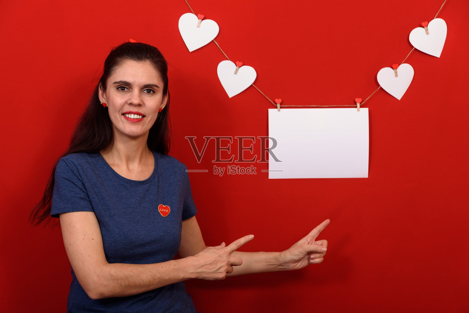 美丽的黑发女人穿着休闲的蓝色t恤在孤立的红色背景附近微笑的矩形横幅和白色的心，并指出这照片摄影图片