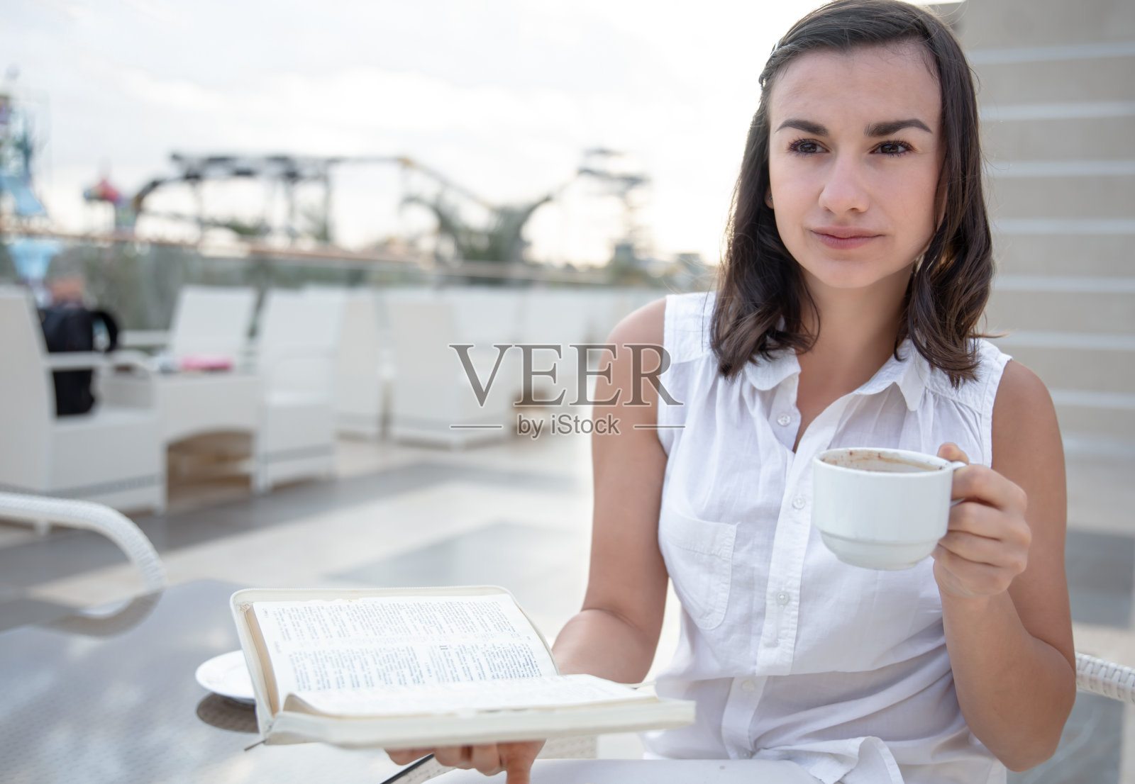 在夏日露台上，手捧咖啡杯和书本的年轻女人照片摄影图片