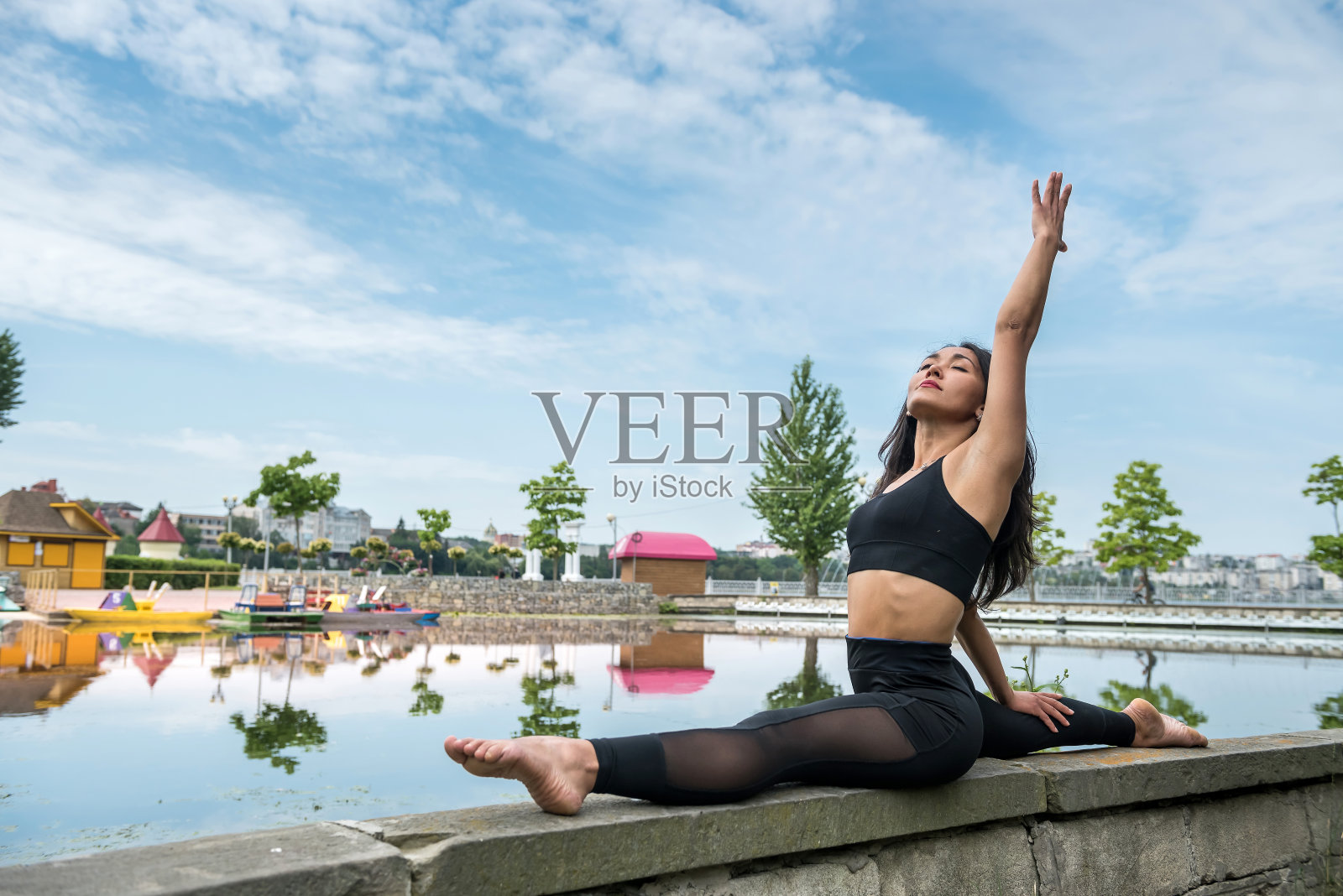 年轻苗条的女人，穿着黑色运动服坐在湖边做伸展运动照片摄影图片