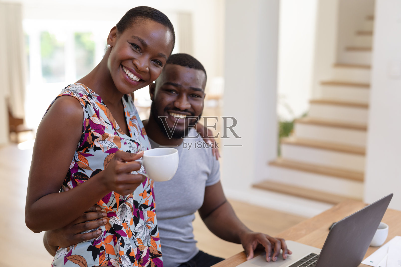 幸福的非裔美国夫妇在厨房里拥抱，用手提电脑的女人端着咖啡照片摄影图片