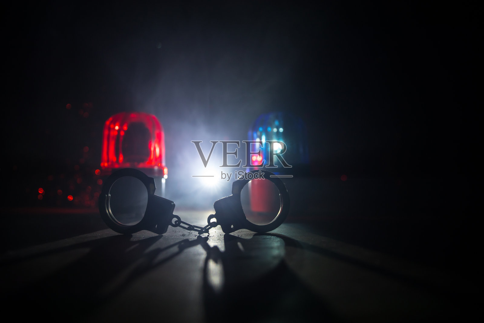 警方夜间突袭，你将被逮捕。手铐的轮廓和警车在后面。在雾蒙蒙的背景下，红色和蓝色警灯闪烁。照片摄影图片