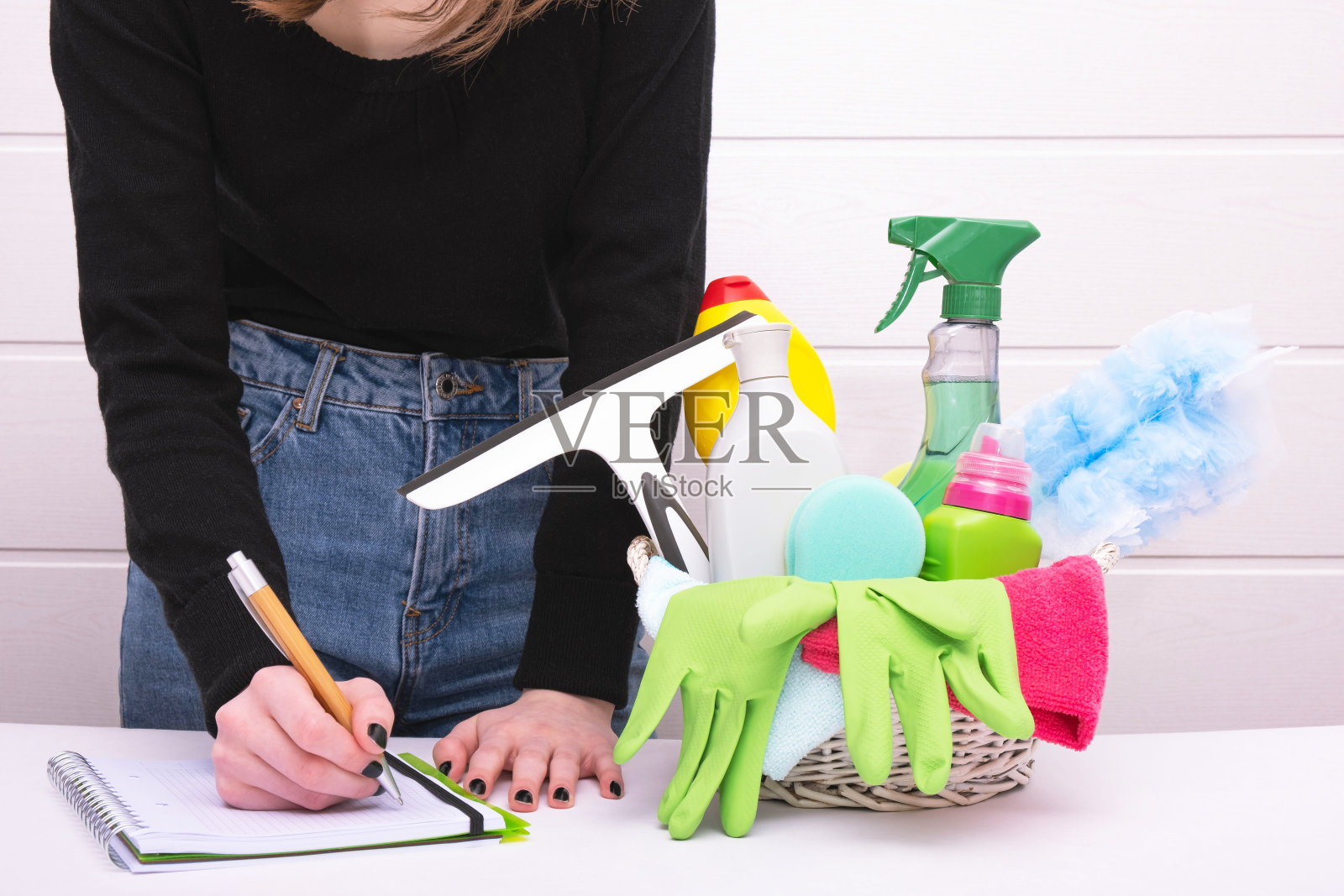 篮子里有清洁用品和消毒剂，用绿色的手套和刷子打扫房子，一个女人在白色的背景上写着一个计划照片摄影图片