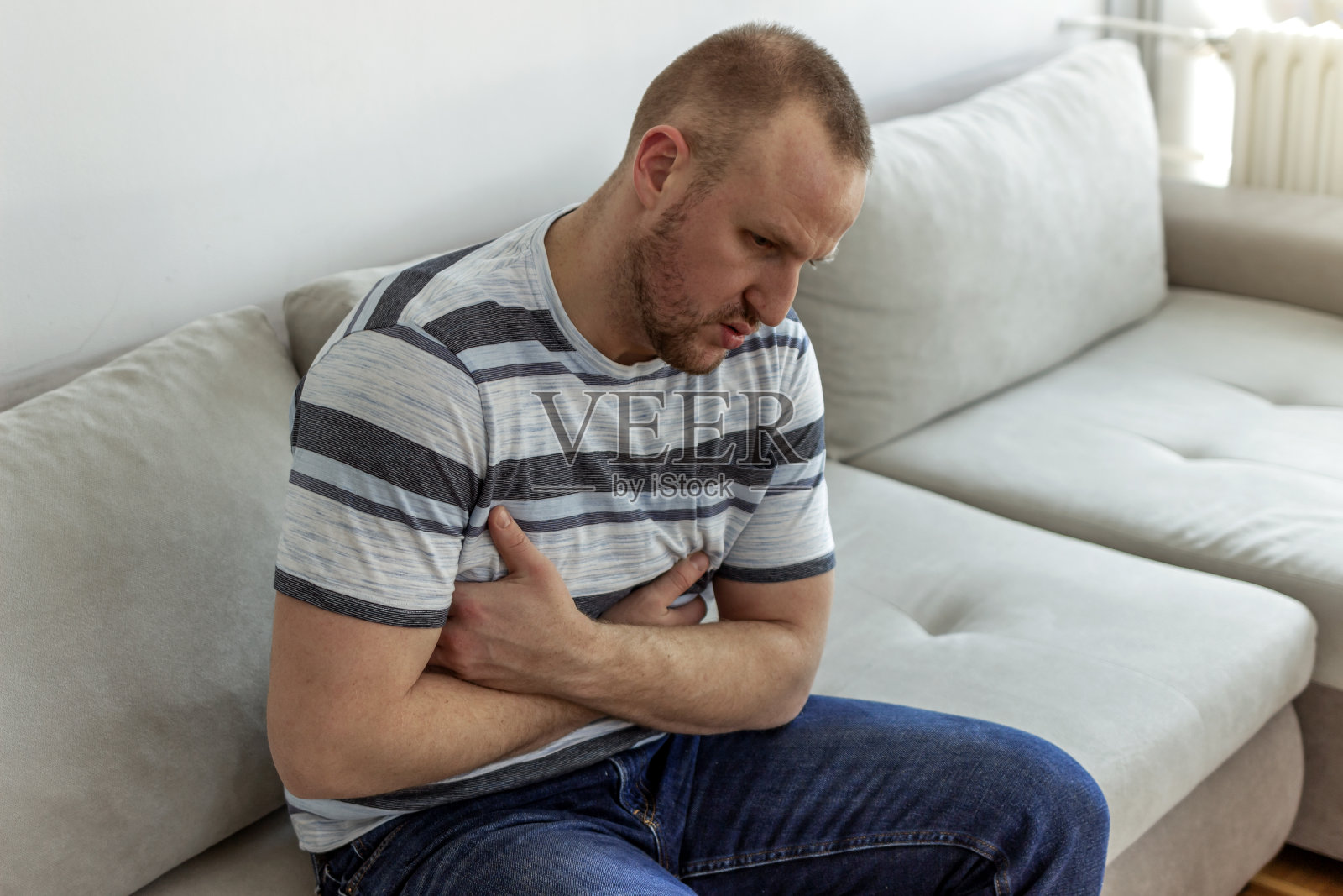 这个男人坐在米色的沙发上，抱着他的肚子。照片摄影图片