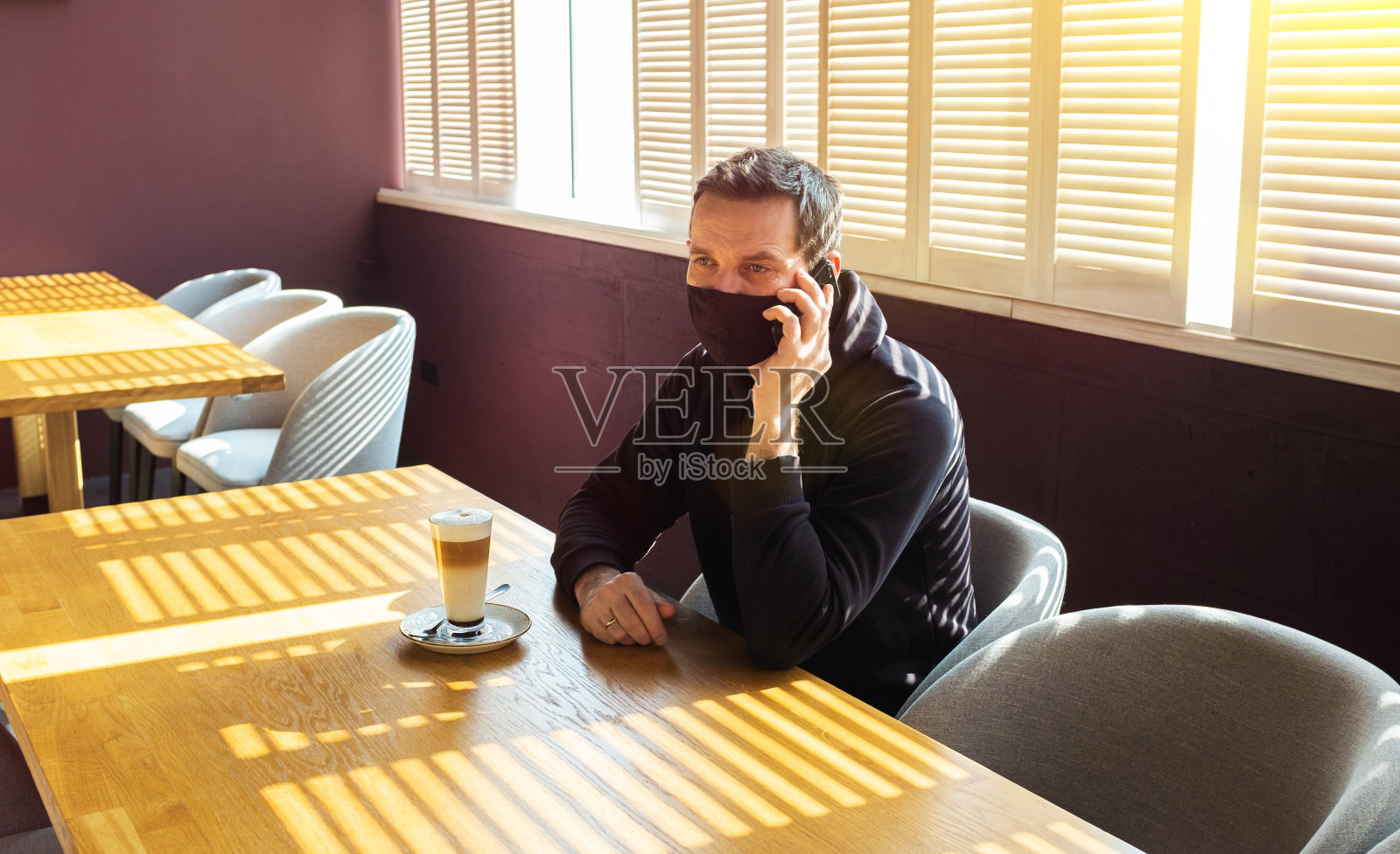 一个白人在咖啡馆里，手里拿着手机。旁边是一杯装在杯子里的卡布奇诺。戴防护口罩，预防冠状病毒感染。新现实的概念照片摄影图片