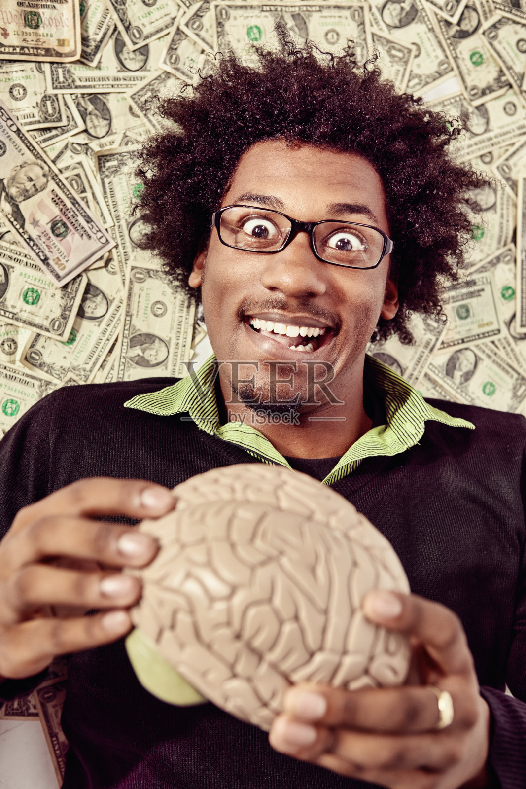 一个戴着眼镜的滑稽、快乐的极客拿着一个人脑模型，背景是大量的现金照片摄影图片