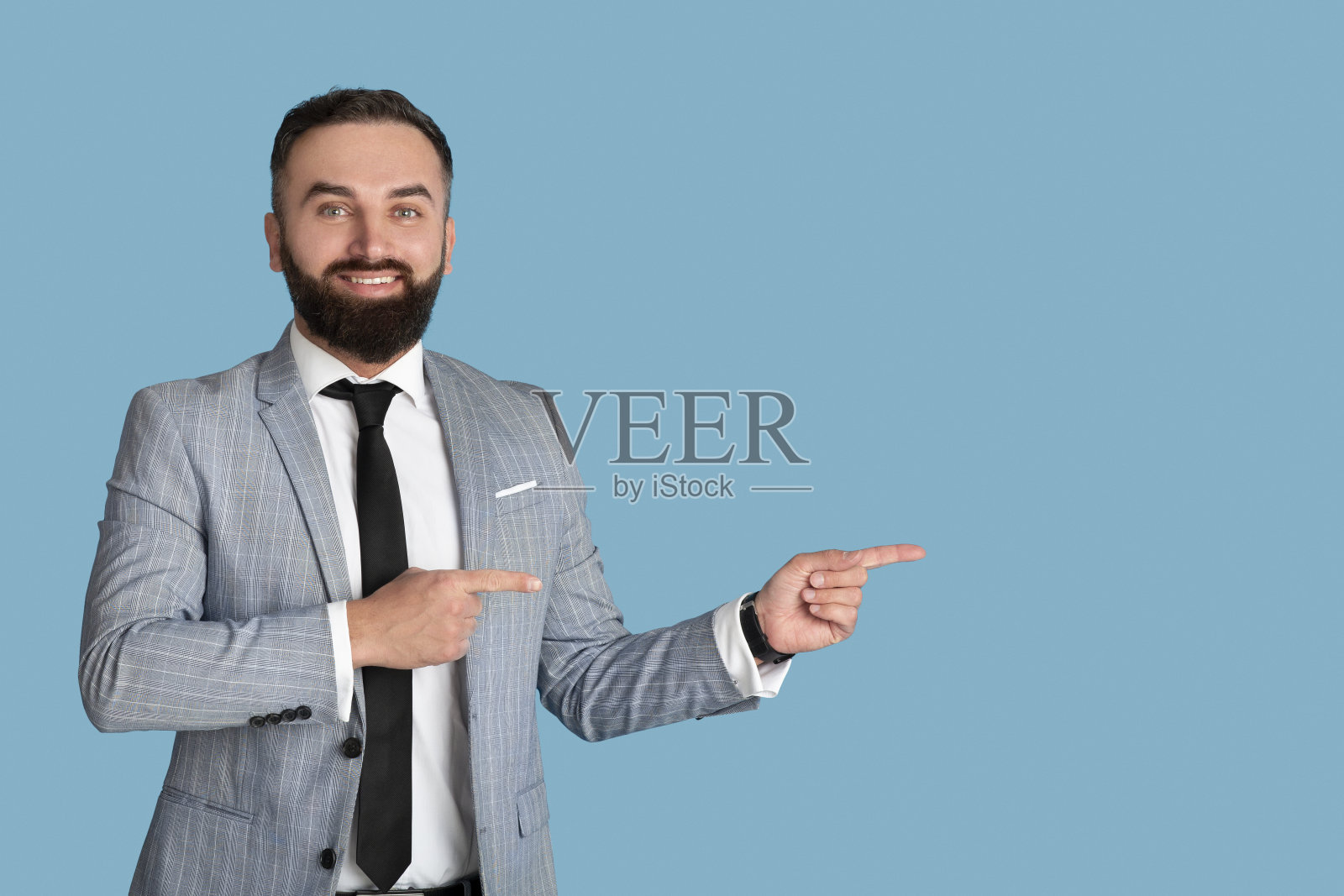 快乐的大胡子商人在正式的西装和领带指向你的广告空间，蓝色的工作室背景照片摄影图片