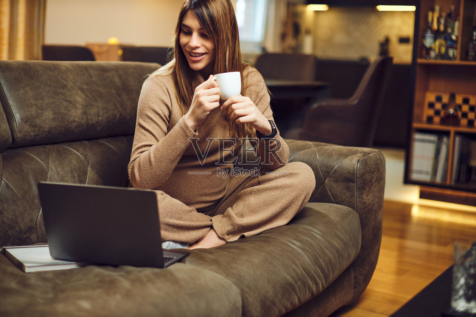 一个孕妇坐在家里的沙发上休息。她一边喝咖啡，一边用笔记本电脑。照片摄影图片