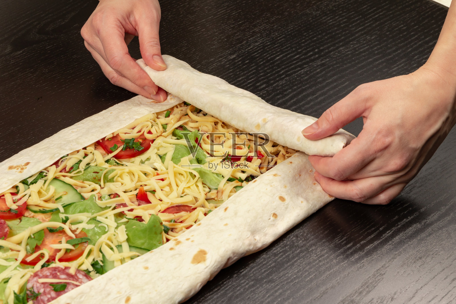 女性用手把新鲜的蔬菜沙拉和奶酪包在皮塔饼里。在木桌上煮沙瓦玛照片摄影图片