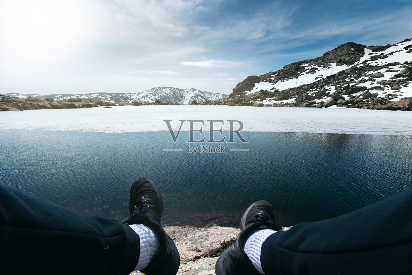 一个男人的脚坐在雪山顶上一个结冰的湖前面。Penalara峰值马德里照片摄影图片
