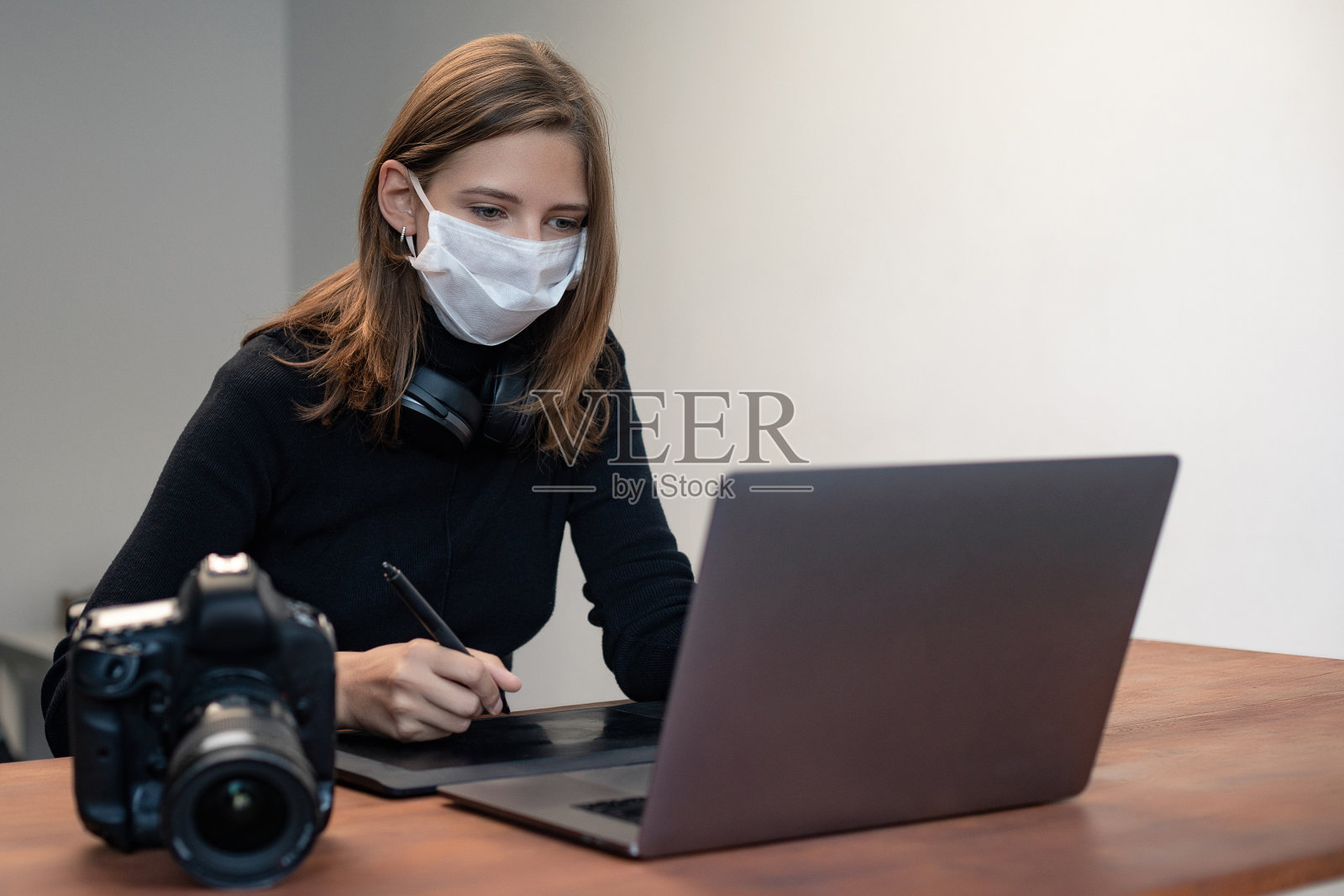 可爱的白人女孩在室内戴医用防护口罩的肖像。戴着无线耳机在笔记本电脑上工作的女人。木制桌子上的专业摄影师数码相机照片摄影图片