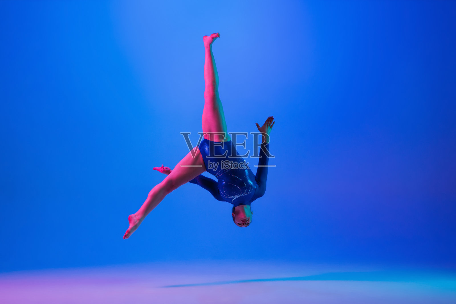 年轻柔顺的女孩孤立在蓝色的工作室背景。年轻的女模特在练习艺术体操。锻炼身体的灵活性和平衡性。照片摄影图片