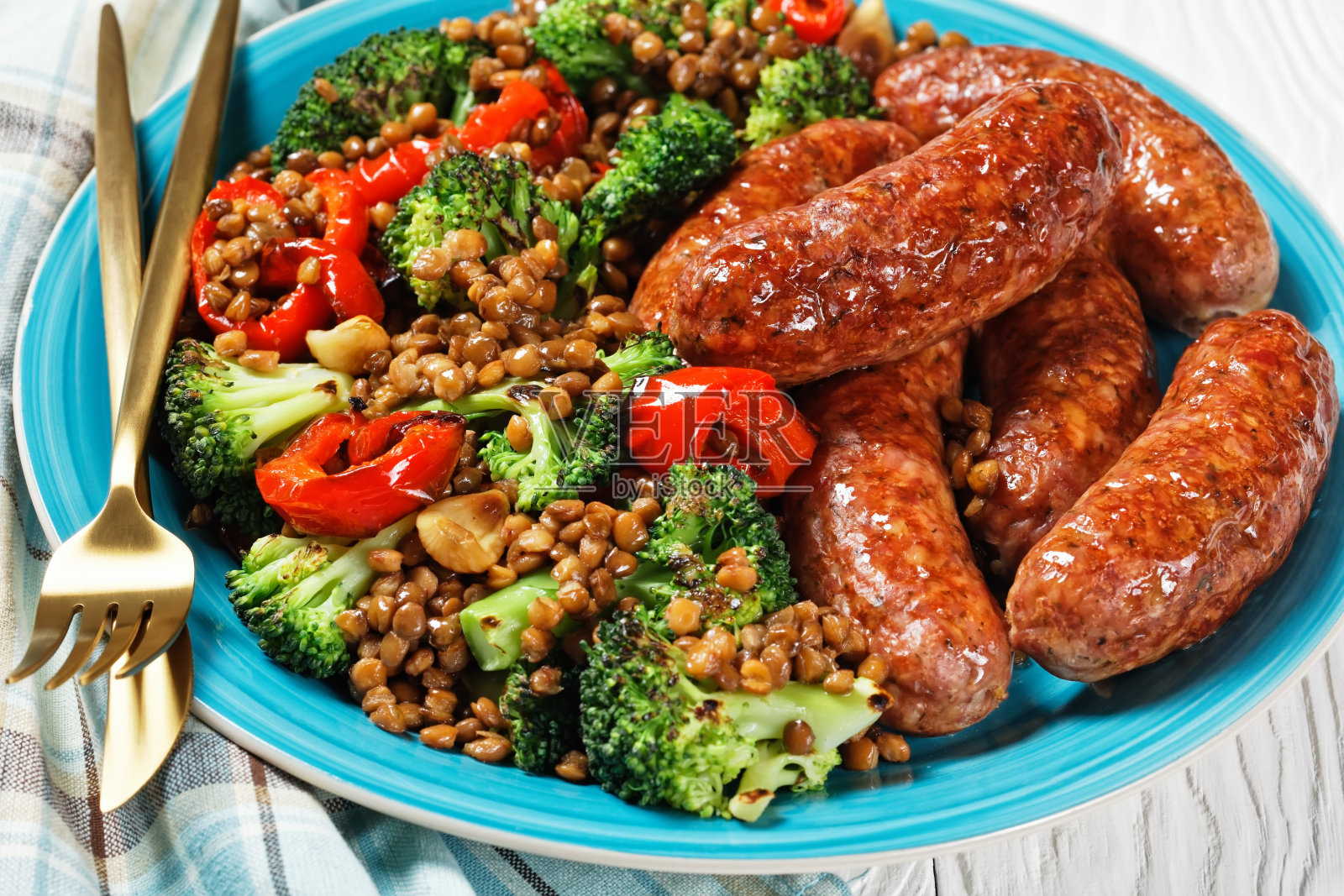 意大利猪肉香肠与红豆、红辣椒和西兰花炖在一个蓝色的盘子里，白色的木质背景上配有金色的餐具、芥末、百里香和番茄酱。俯视图，特写照片摄影图片