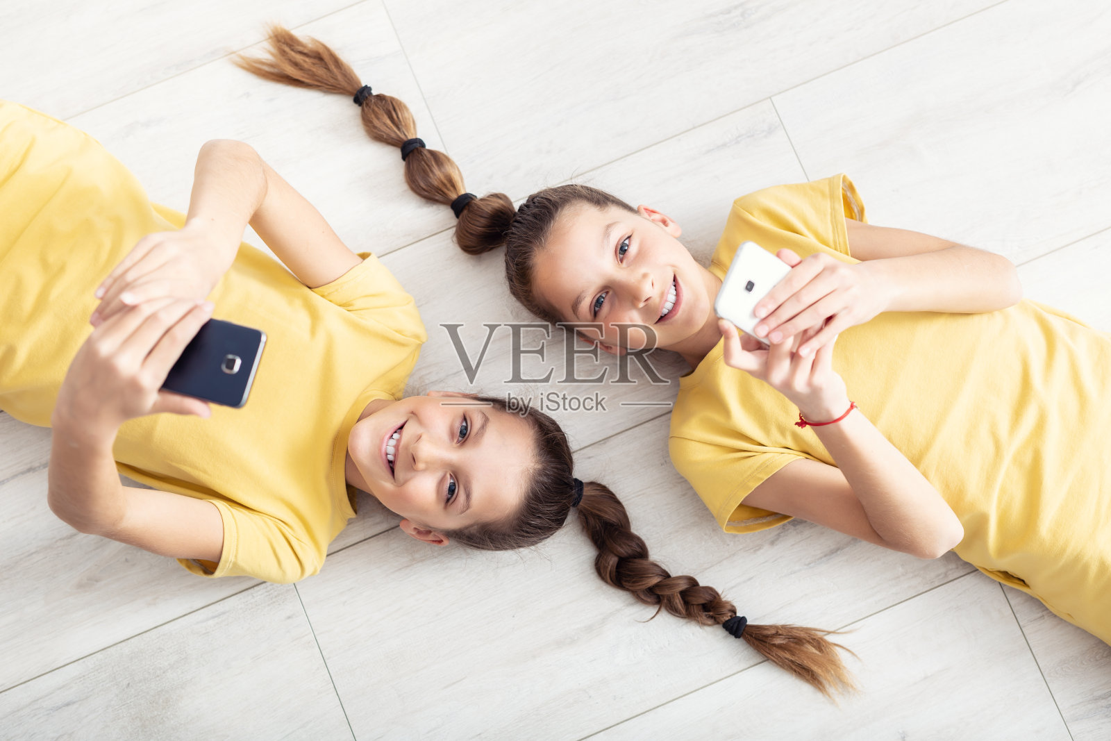一对年轻的双胞胎女孩躺在地板上，手里都拿着手机。照片摄影图片