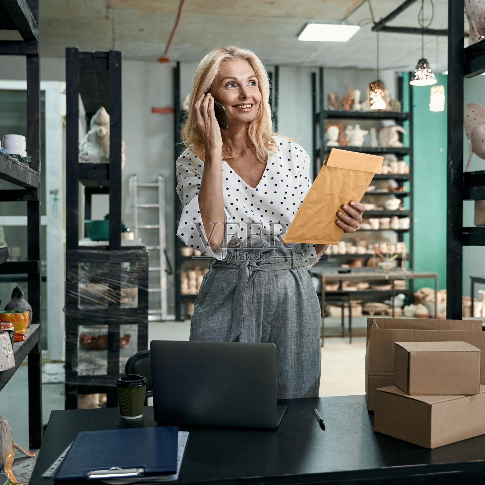 年轻成功的女企业主一边拿着信封一边打电话，一边在艺术工作室或陶艺店用手工陶瓷制品工作照片摄影图片