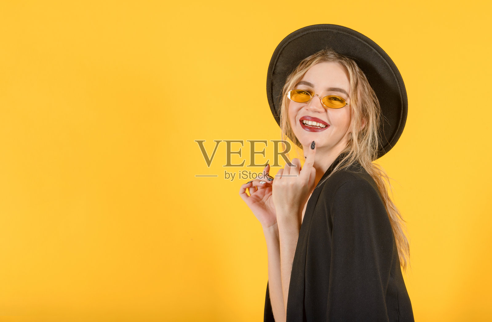 白种年轻的厚脸皮金发女人穿着黑色的西装，帽子和眼镜在黄色的背景微笑照片摄影图片