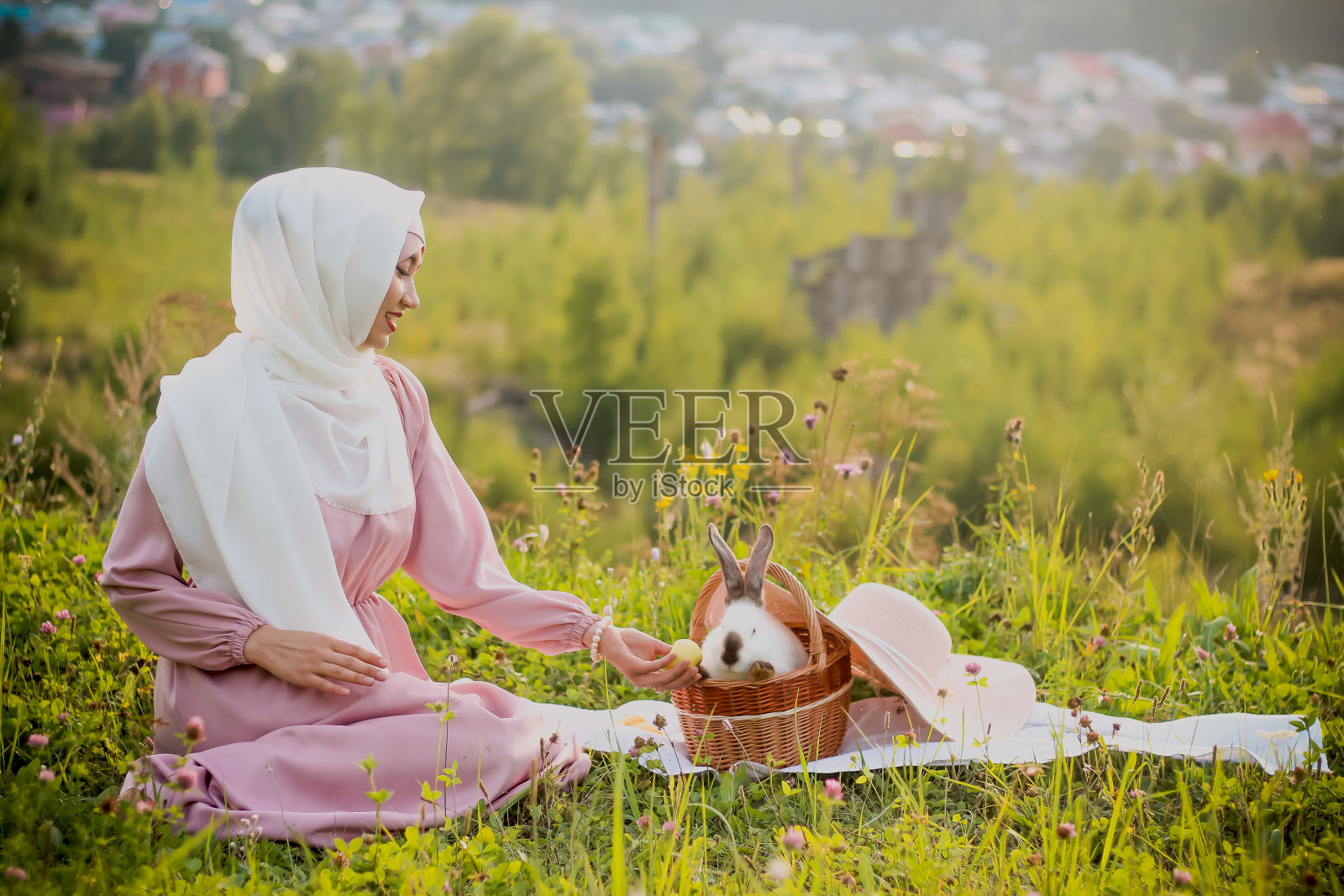 一名穆斯林妇女坐在球场上，旁边是一个篮子，篮子里放着一只兔子和一顶帽子。一个穆斯林妇女的肖像戴着粉色的头巾，珍珠手镯。温柔。照片摄影图片
